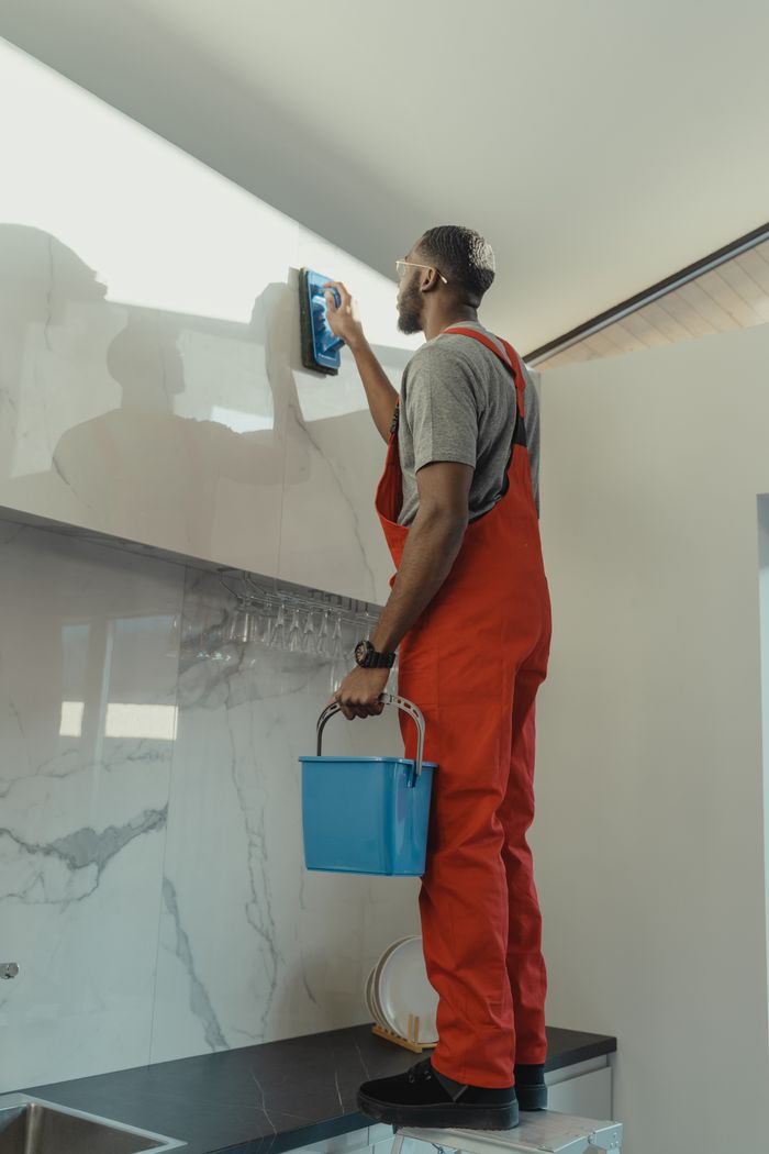 services-03 African American man in red overalls cleaning a kitchen wall with a brush and pail.
